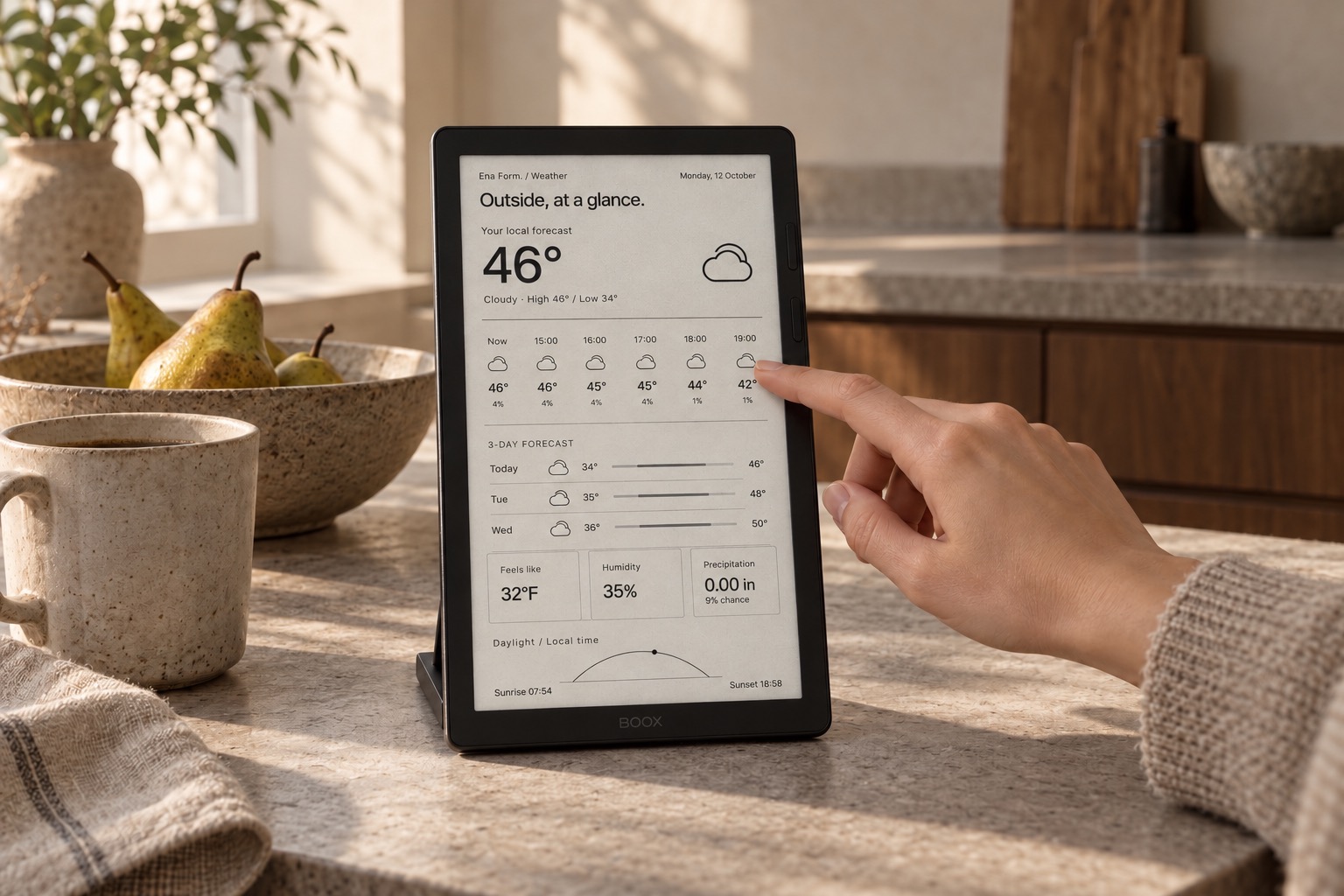 A small e-ink display showing a monochrome weather view beside ceramics on a sunlit kitchen counter, with someone selecting the forecast.